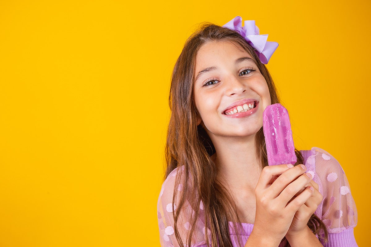 Menina sorridente segurando um picolé de amora em frente a um fundo amarelo vibrante
